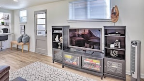 A living room with a large flat-screen TV, a dark wooden entertainment unit with glass doors, a woven bench, and a radiator. - Cozy Rapid City Retreat: Perfect for Exploring! (Rapid City)