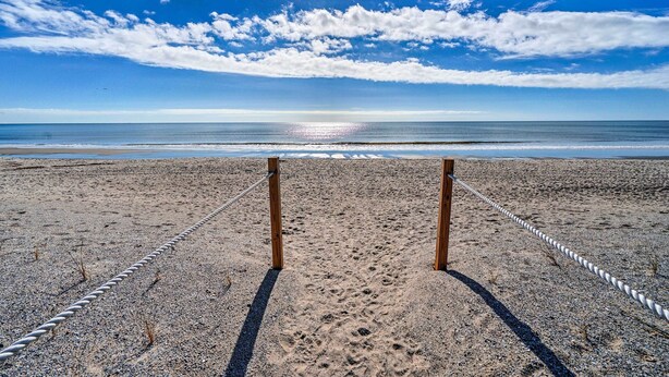 A beach with a rope barrier and wooden posts, leading to the ocean. - Oak Island Beachfront Condo w/ Community Pool (Oak Island)