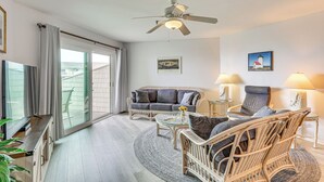 A spacious living room with a large sofa, two armchairs, a coffee table, and a ceiling fan. There are two side tables with lamps, a large window with curtains, and a painting on the wall. - Oak Island Beachfront Condo w/ Community Pool (Oak Island)