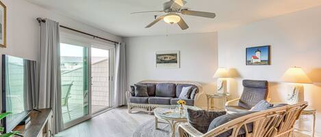 A spacious living room with a large sofa, two armchairs, a coffee table, and a ceiling fan. There are two side tables with lamps, a large window with curtains, and a painting on the wall.
