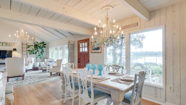 A dining area with a wooden table, white chairs, and a chandelier. There is a large window with a view of the outdoors.