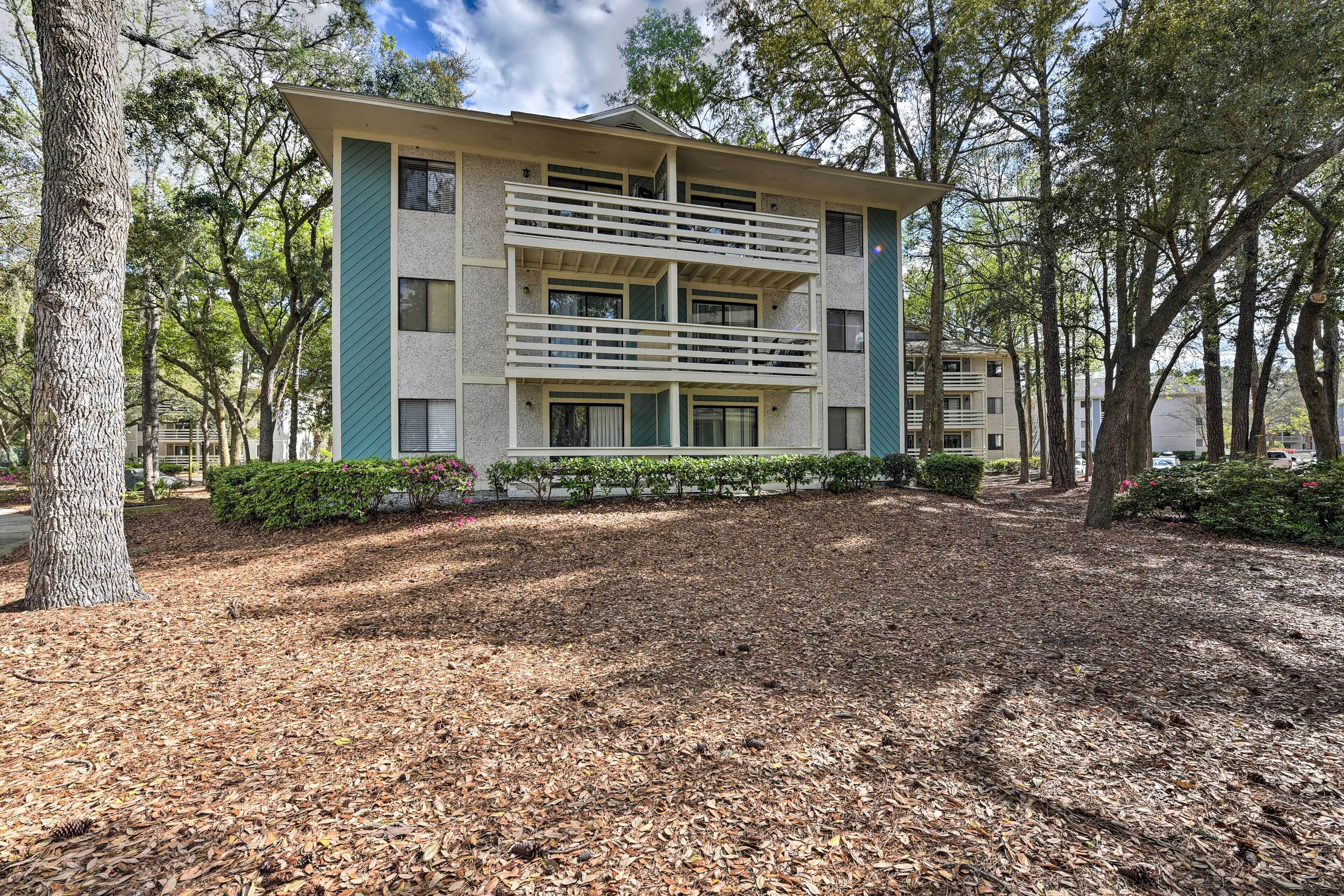 A multi-story building with a balcony on the second floor, surrounded by trees and a leaf-covered ground.