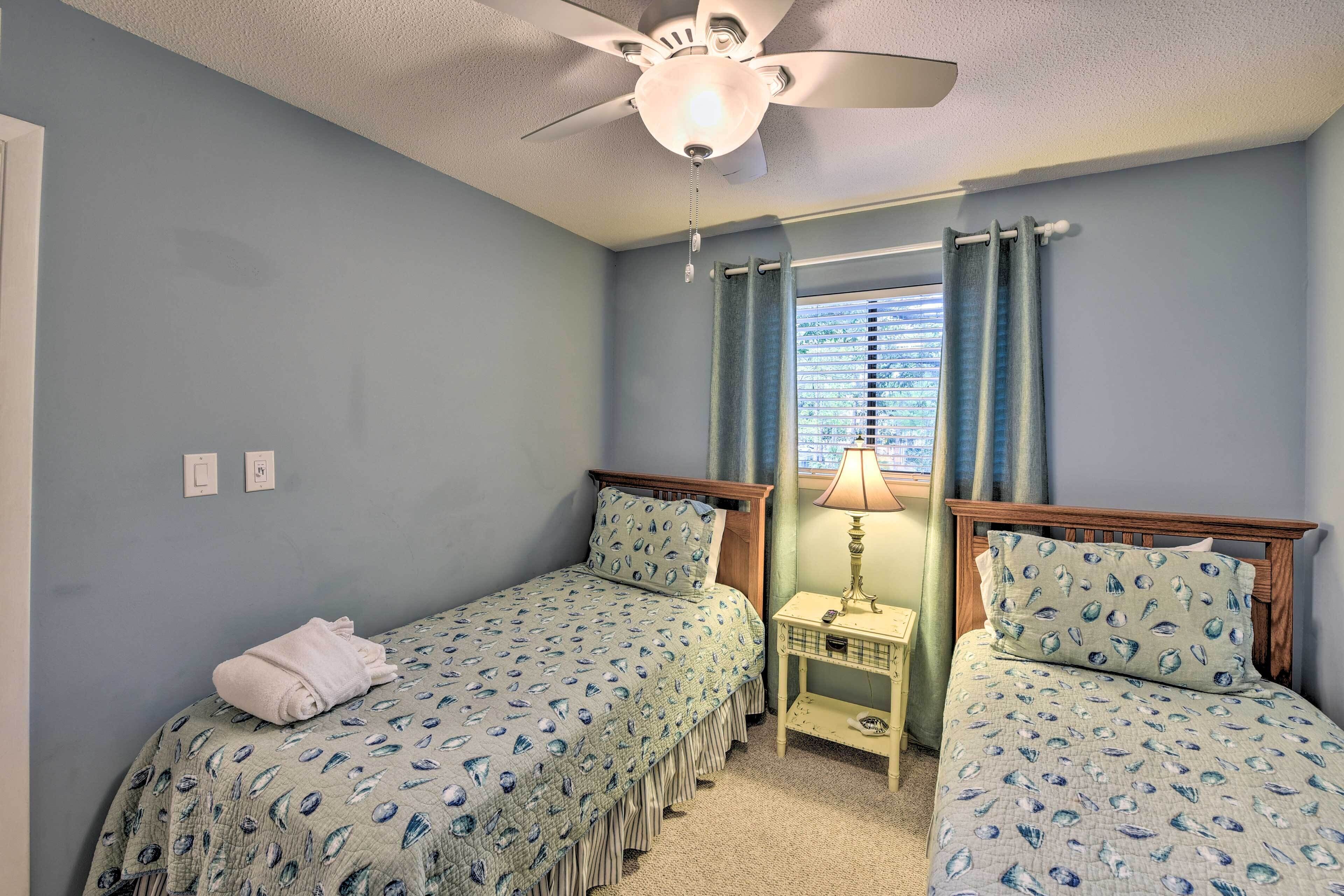A bedroom with two single beds, a ceiling fan, a window with curtains, and a bedside table with a lamp.