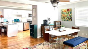 Een moderne keuken met witte kastjes, een eethoek met een witte tafel en bruine stoelen, een plafondventilator en een kleurrijk vloerkleed.