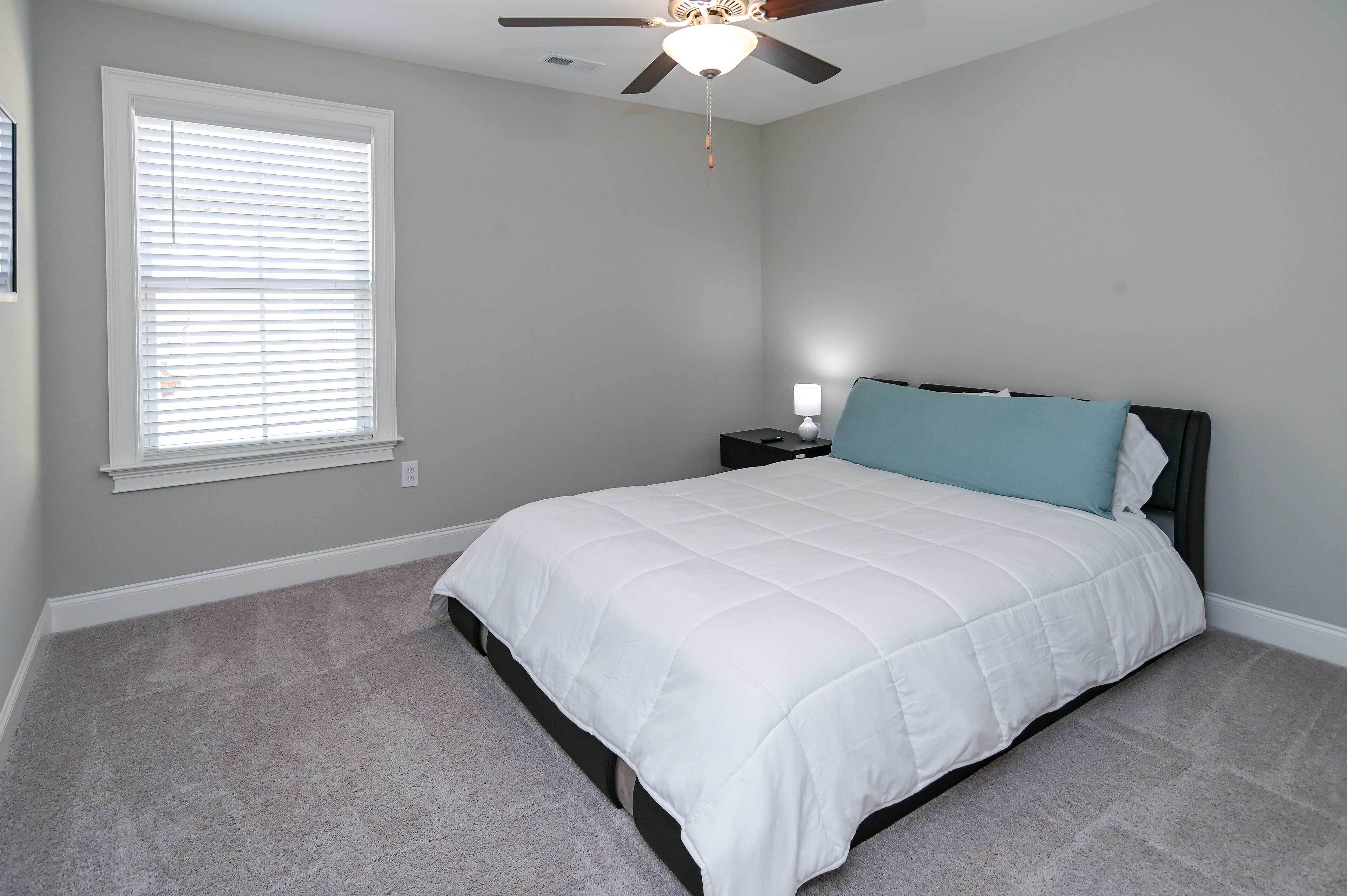 A bedroom with a bed, a ceiling fan, a window with blinds, and a nightstand with a lamp.