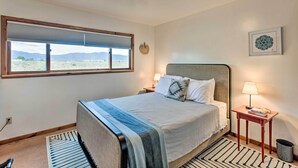 A bedroom with a large window, a bed with a blue and white bedspread, a bedside table with a lamp, and a rug. - El Prado Duplex w/ Deck + Grill - 4 Mi to Town! (Taos)