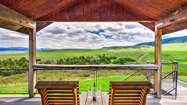 Una mesa y sillas de madera en un porche cubierto, con vistas a un paisaje verde y montañas al fondo.