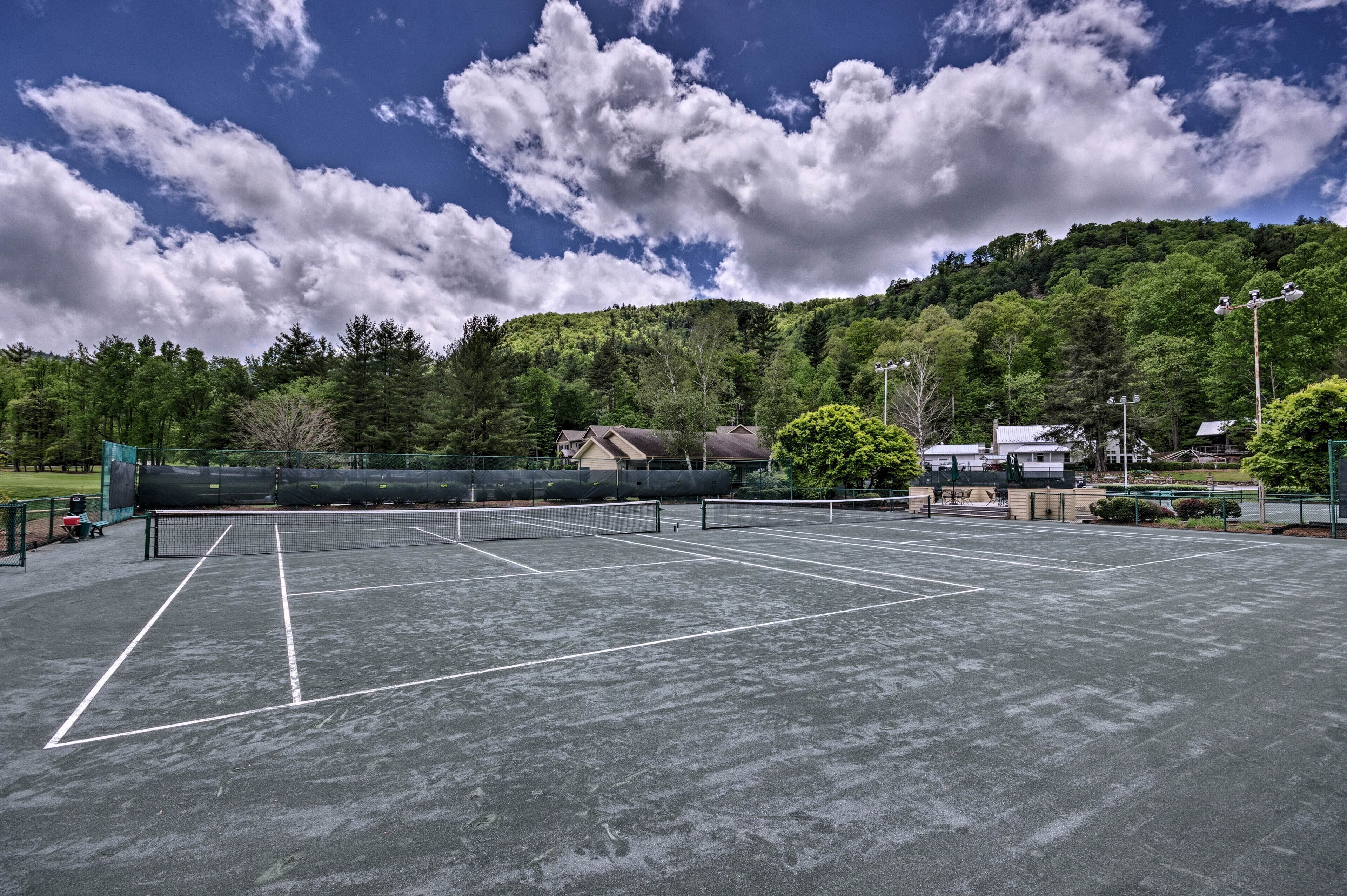 A tennis court with a net, surrounded by trees and a hill in the background.