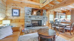 A cozy log cabin interior with a stone fireplace, wooden furniture, and a dining area.