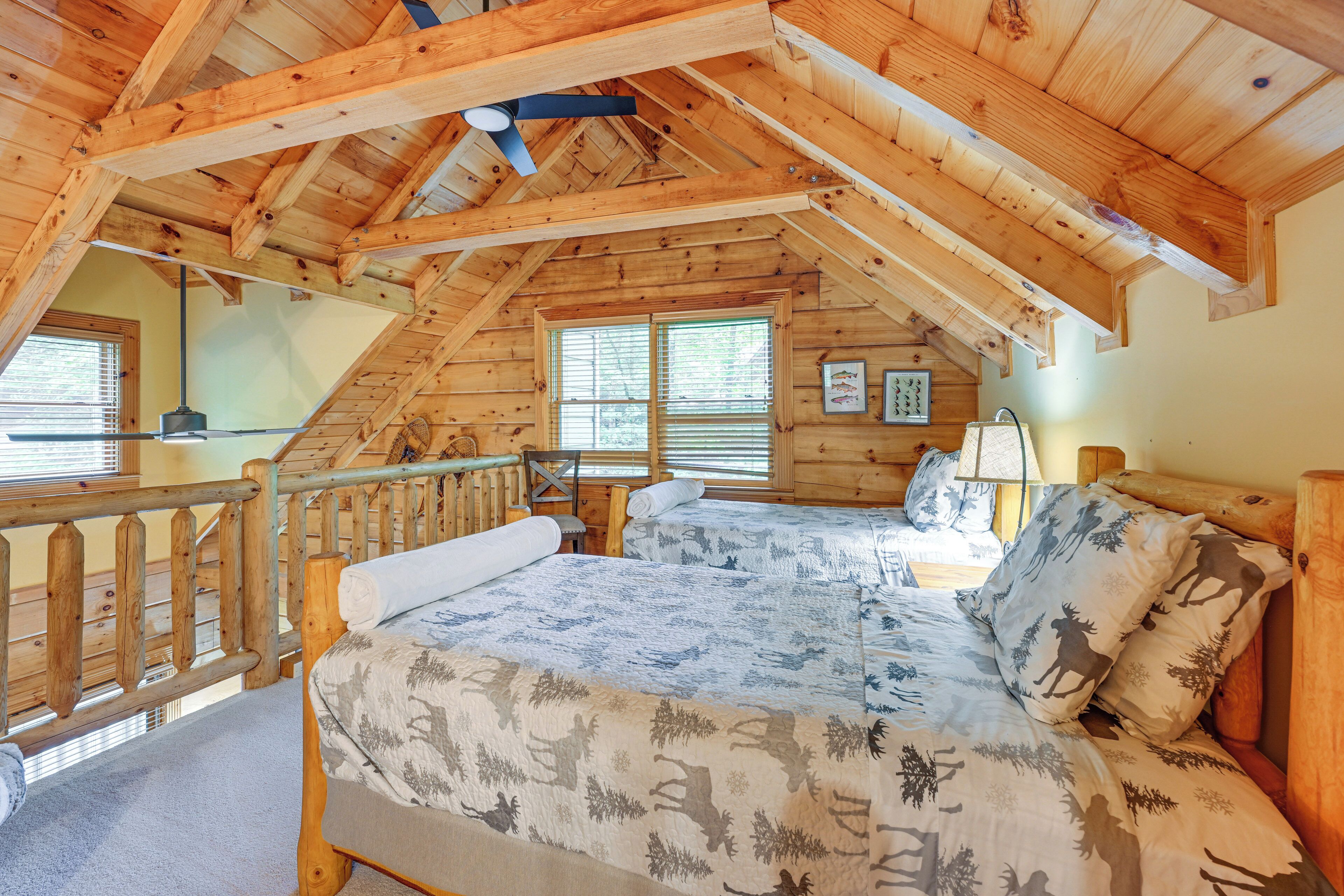 Una cabaña de madera con habitación superior que cuenta con una cama con ropa de cama estampada, un ventilador de techo y vistas al exterior a través de una ventana.