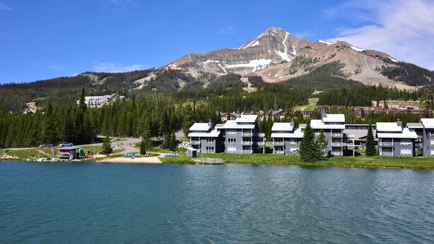 Un complejo turístico junto al lago con edificios modernos, montañas al fondo y cielo despejado.