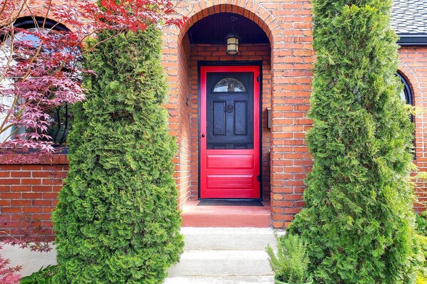 A red door with a black frame, flanked by two evergreen shrubs, leading to a brick entrance.