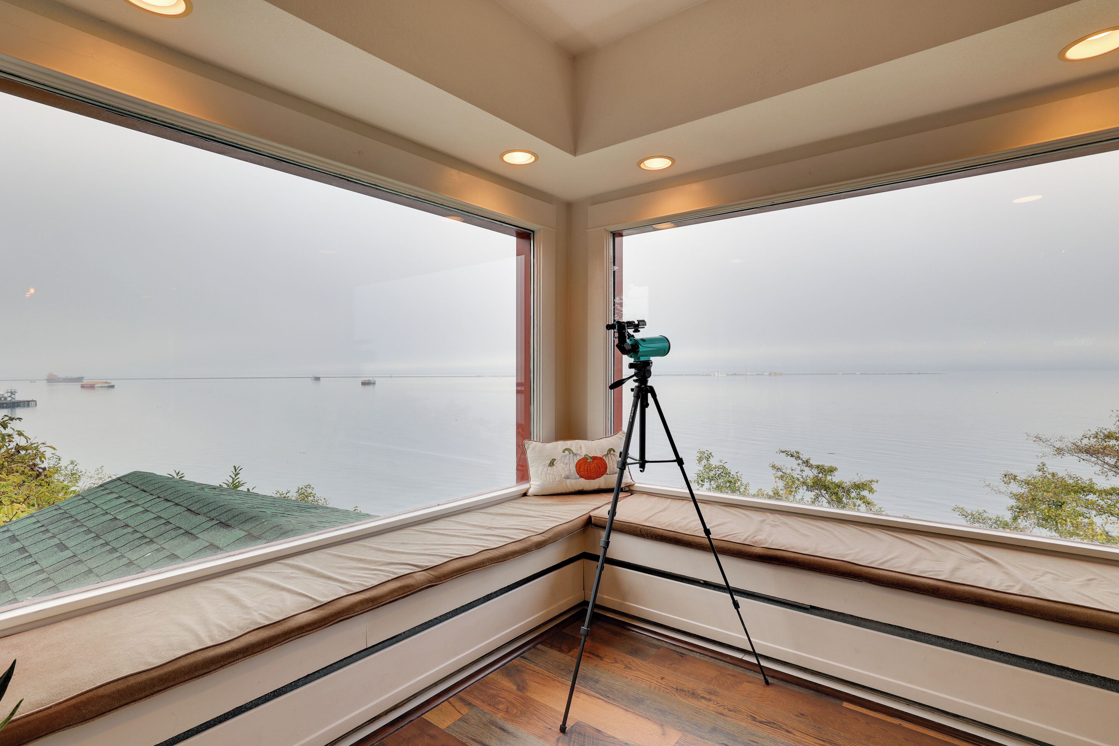 A room with large windows overlooking a body of water, a telescope on a tripod, and a cushioned bench with a pillow.