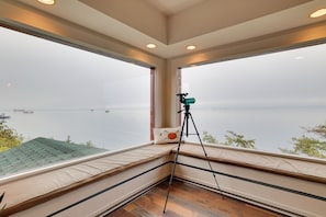 A room with large windows overlooking a body of water, a telescope on a tripod, and a cushioned bench with a pillow. - Chic Port Angeles Home w/ Oceanfront Balcony! (Port Angeles)