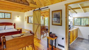 A wooden cabin interior with a bed, desk, rocking chair, and a kitchen area. - Secluded Tallassee Cabin w/ Fire Pit & Porch! (Tallassee)