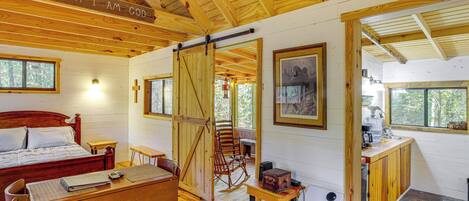 A wooden cabin interior with a bed, desk, rocking chair, and a kitchen area.