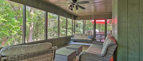 A covered porch with wicker furniture, a ceiling fan, and large windows overlooking a wooded area.