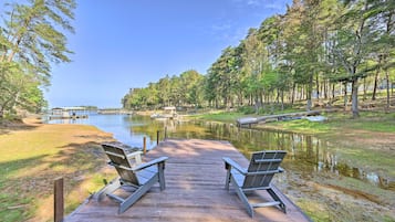 A wooden dock with two chairs facing a calm lake, surrounded by trees and a distant building.