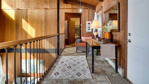 Un interior de madera con una escalera, un tapete estampado, una silla de mimbre, una mesa con un florero y un espejo en la pared.