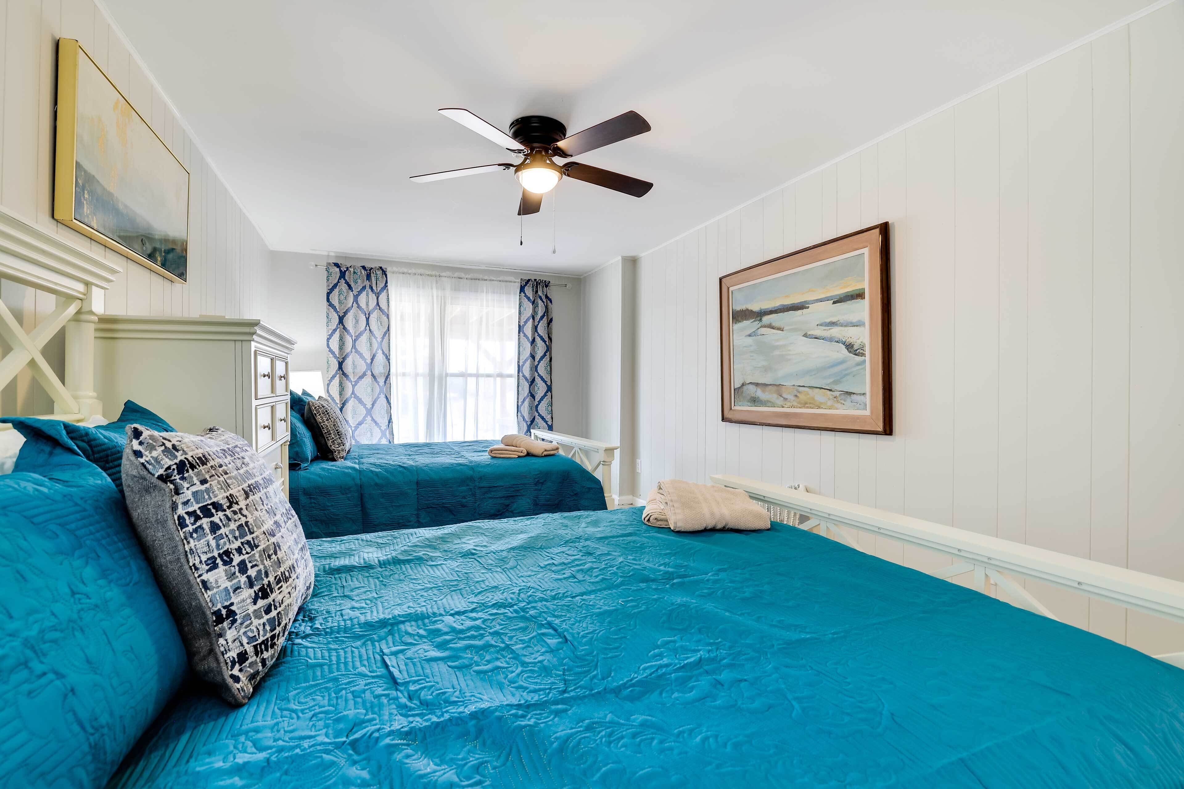 A bedroom with two beds, a ceiling fan, a window with patterned curtains, and a painting on the wall.
