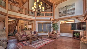 A spacious living room with a high ceiling, wooden beams, a chandelier, leather sofas, a flat-screen TV, and a staircase leading to an upper level. - Grand Angel Fire Retreat w/ Hot Tub & Fire Pit! (Angel Fire)