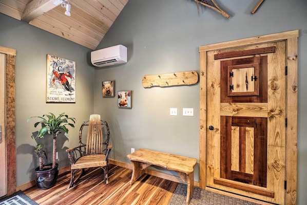A room with a wooden door, a bench, a chair, a potted plant, and wall decorations including a vintage poster.