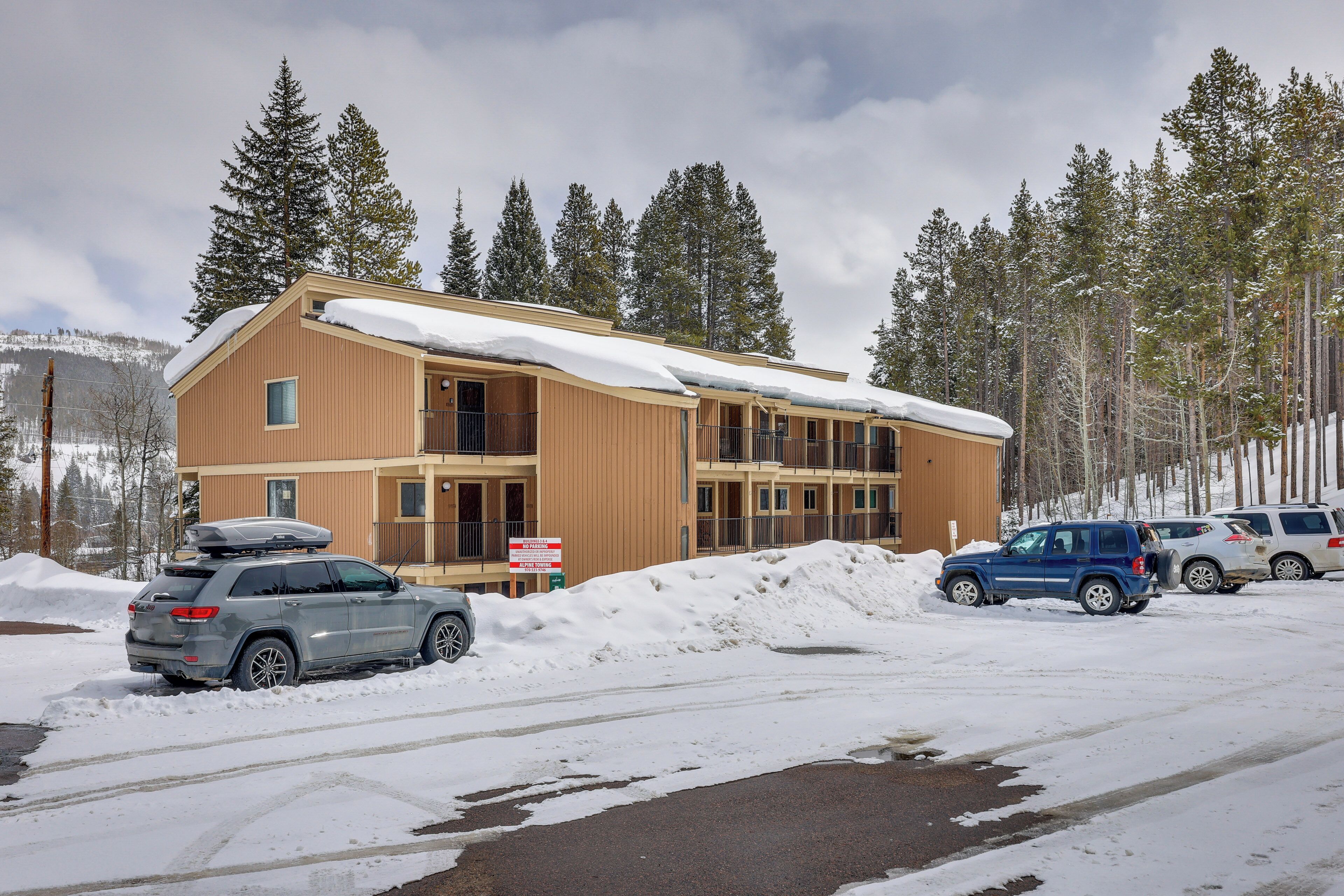 A multi-story building with a snow-covered roof, surrounded by tall pine trees and a mountainous backdrop.