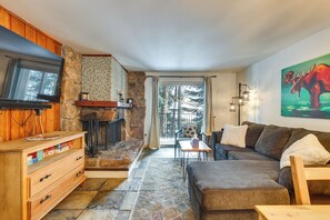 A cozy living room with a stone fireplace, a large flat-screen TV, a sofa, and a painting of a moose. - Winter Park Condo Rental ~ 2 Mi to Ski Resort! (Winter Park)