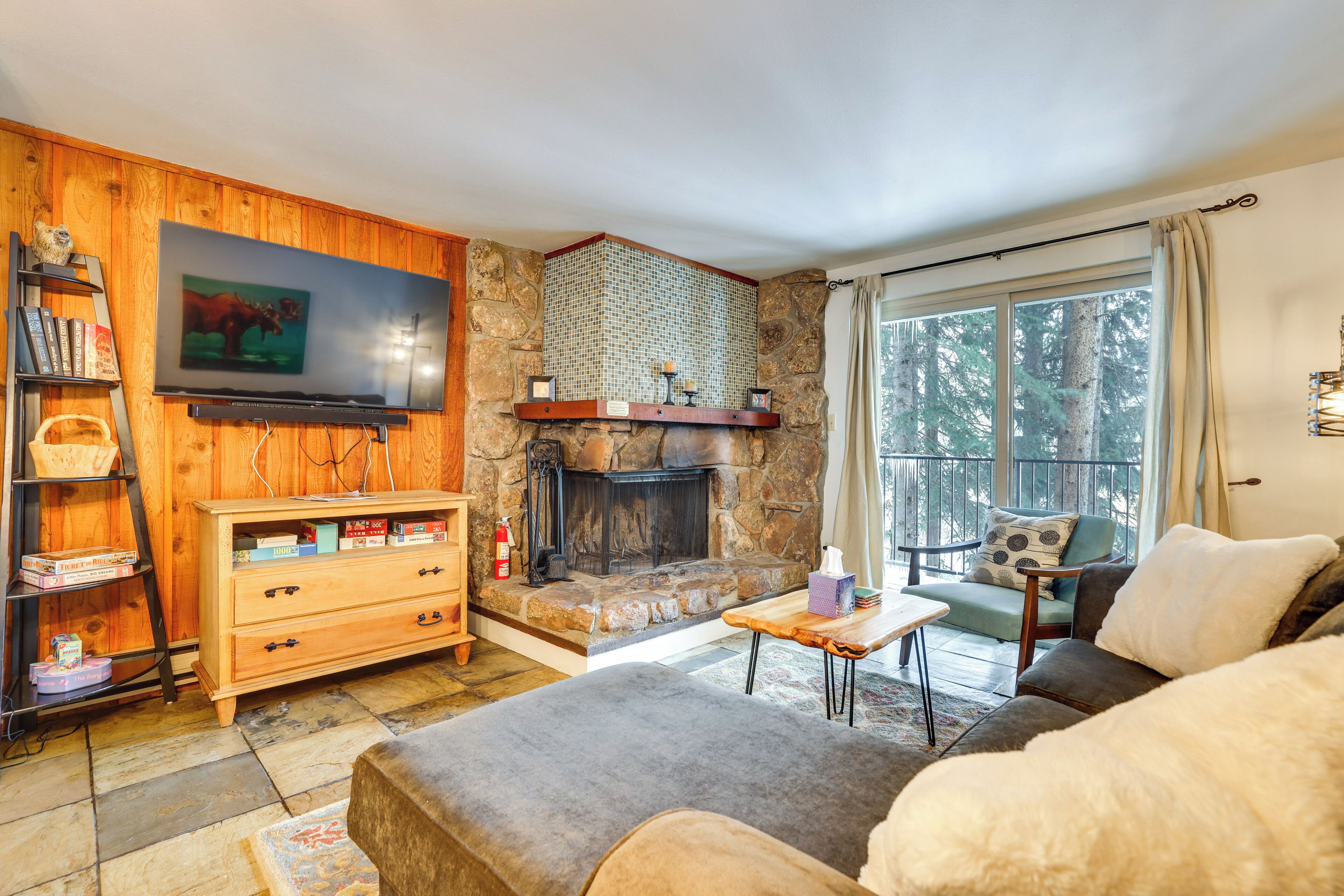 Ein gemütliches Wohnzimmer mit Kamin aus Stein, Holzregalen, einem Fernseher und Blick auf Bäume durch ein großes Fenster.