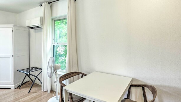 Quarto com uma mesa branca, duas cadeiras, um ventilador, um varal, uma janela com cortinas e um aparelho de ar condicionado.
