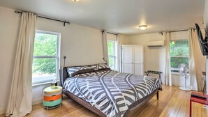 Quarto com cama, mesa lateral redonda, janela com cortinas, ar condicionado e ventoinha.