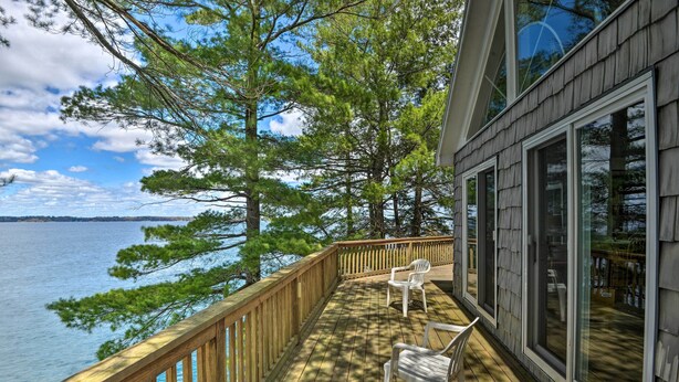 A wooden deck with a railing overlooking a body of water, two white chairs, and large windows.