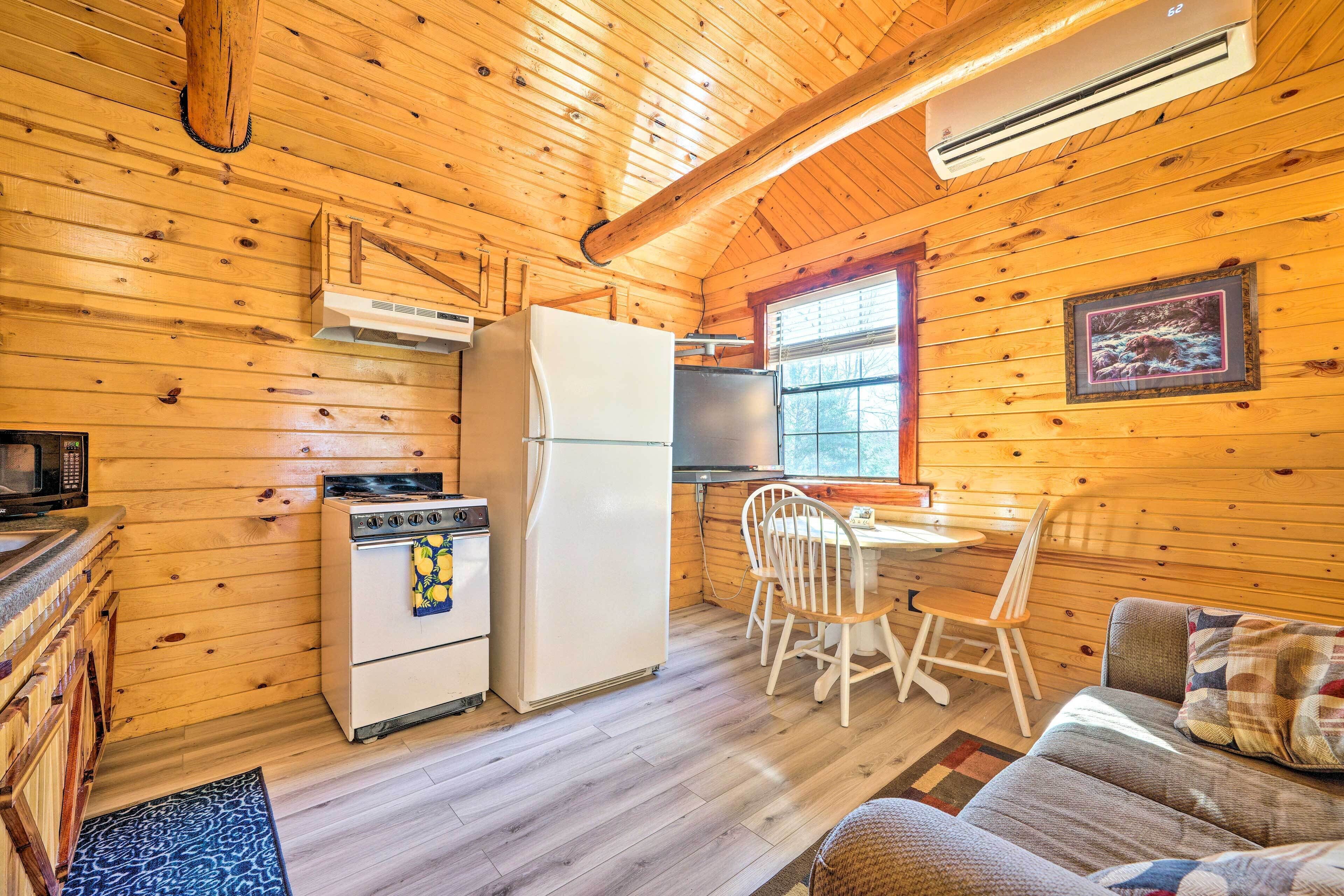 A cozy cabin interior with wooden walls, a kitchen area including a refrigerator, stove, and microwave, a dining table with chairs, and a sofa with a patterned cushion.