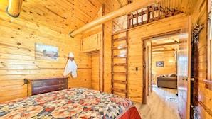 Interior de una cabaña de madera con una cama, una escalera de madera y una puerta que da a otra habitación.