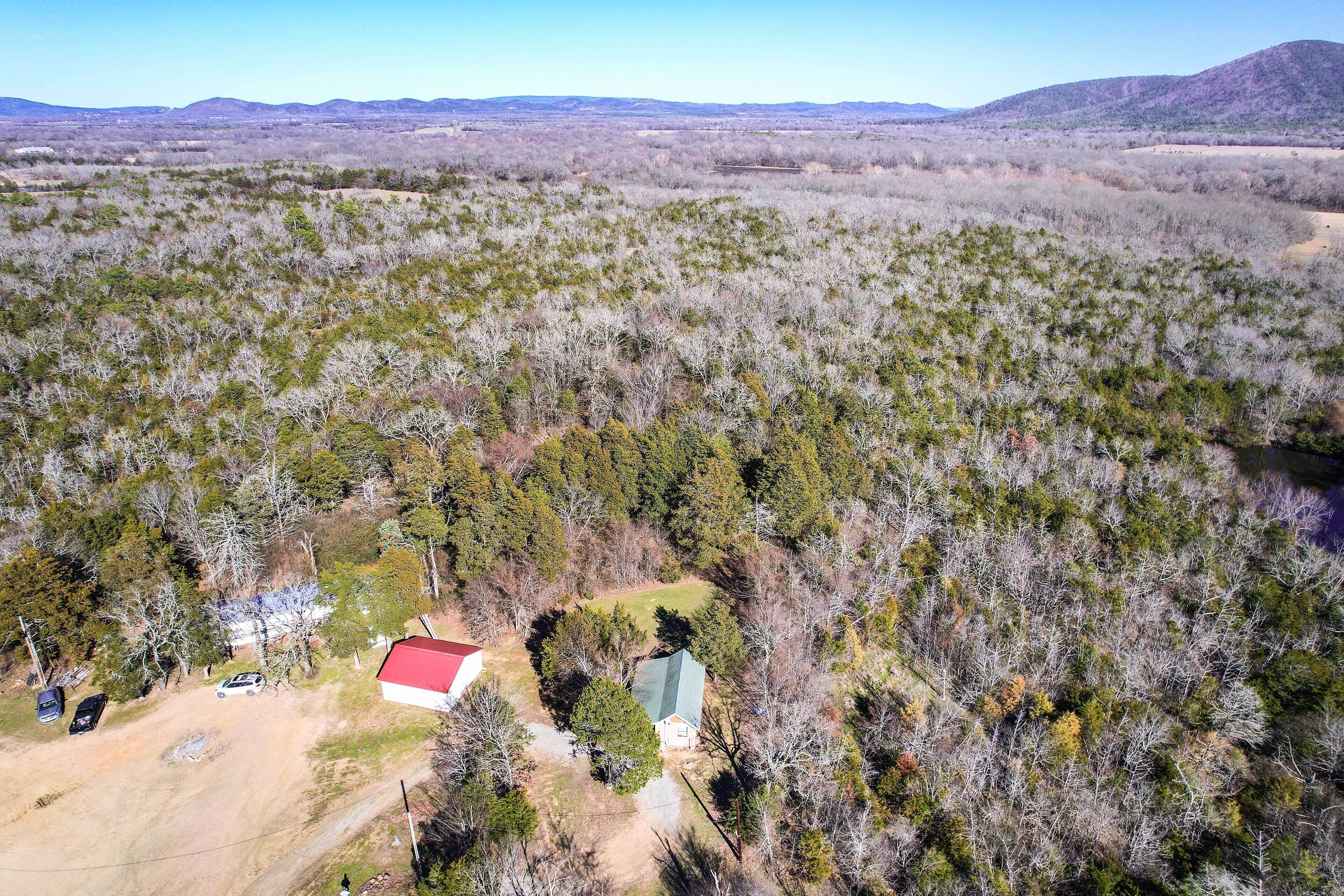 Vue aérienne d’une propriété rurale avec une maison entourée d’une forêt dense, une grange au toit rouge et un chemin de terre.