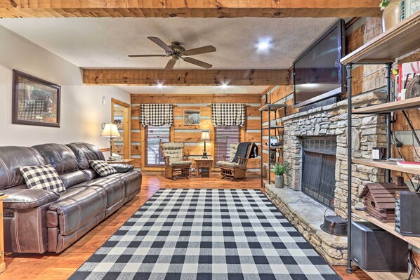 A cozy living room with a checkered rug, leather sofa, wooden walls, a stone fireplace, and a ceiling fan.