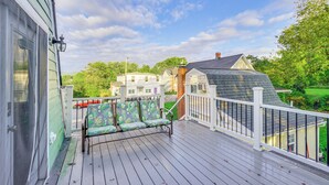 A wooden deck with a white railing, two green and white patterned chairs, and a view of a house with a green roof and a brick chimney. - Epic Brunswick Apartment With Hot Tub! (Brunswick)