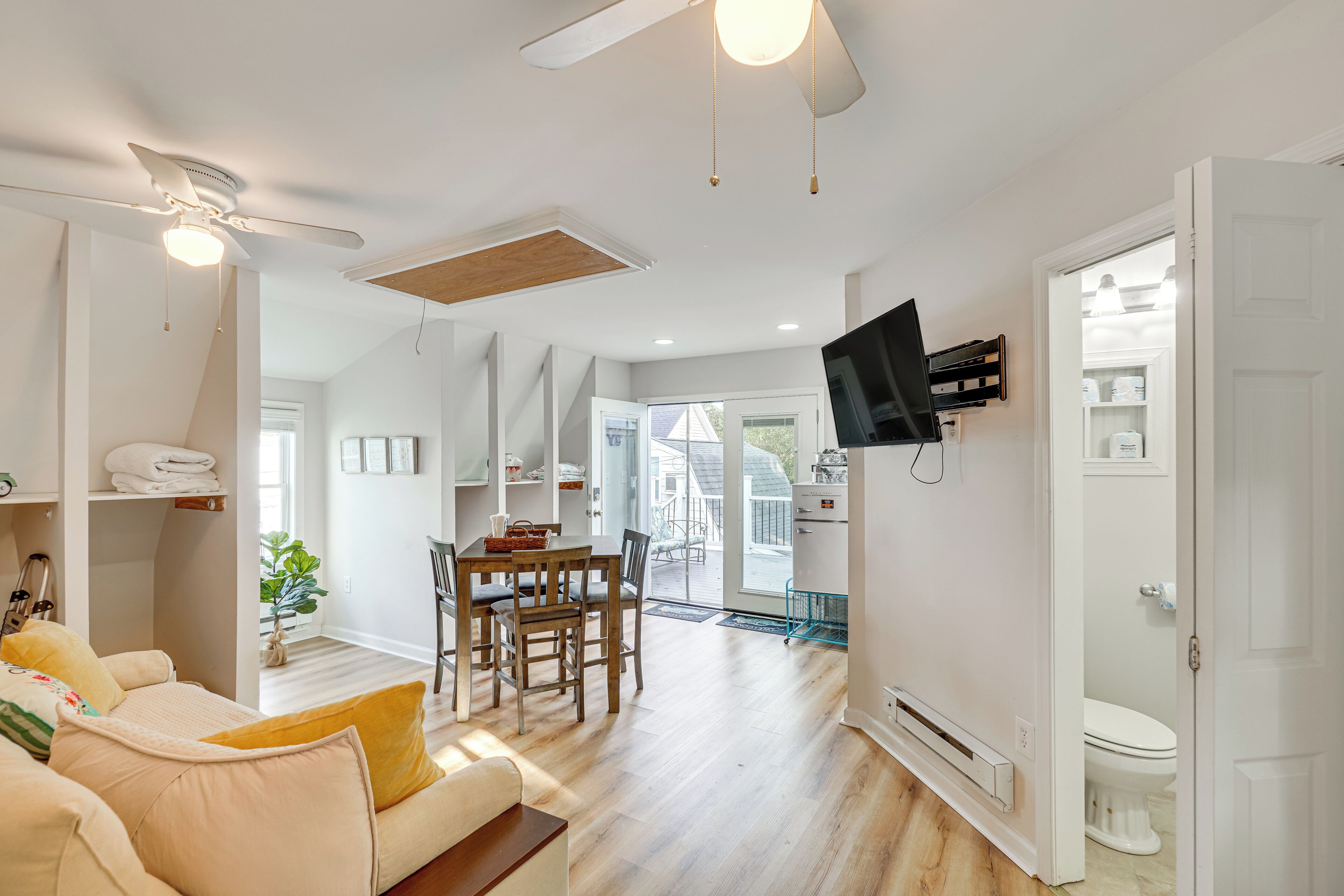 A modern living room with a sofa, dining table, and chairs. There is a ceiling fan, a TV mounted on the wall, and a door leading to a bathroom.