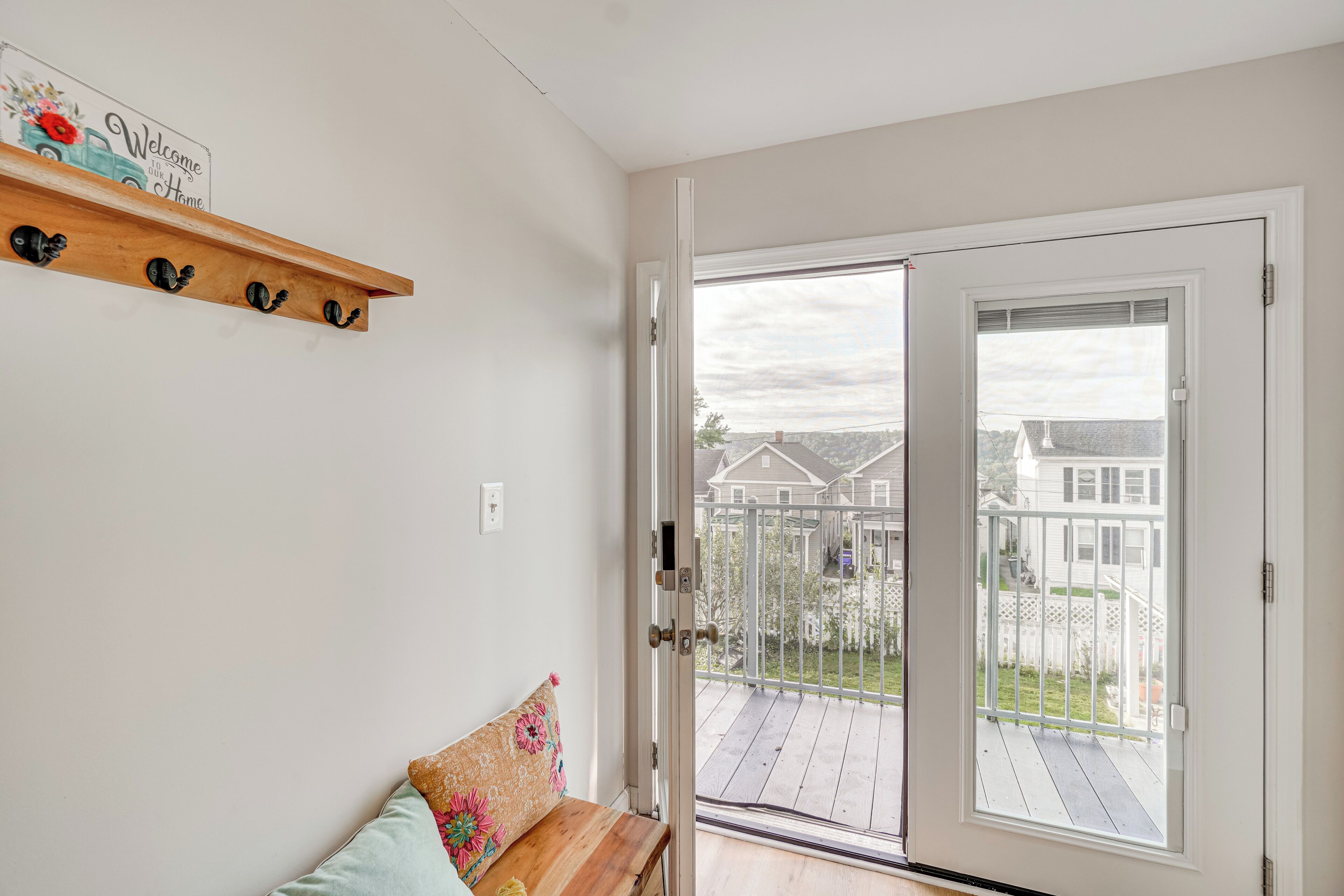 A room with a wooden bench, a welcome sign, and a sliding glass door leading to a balcony with a view of houses.