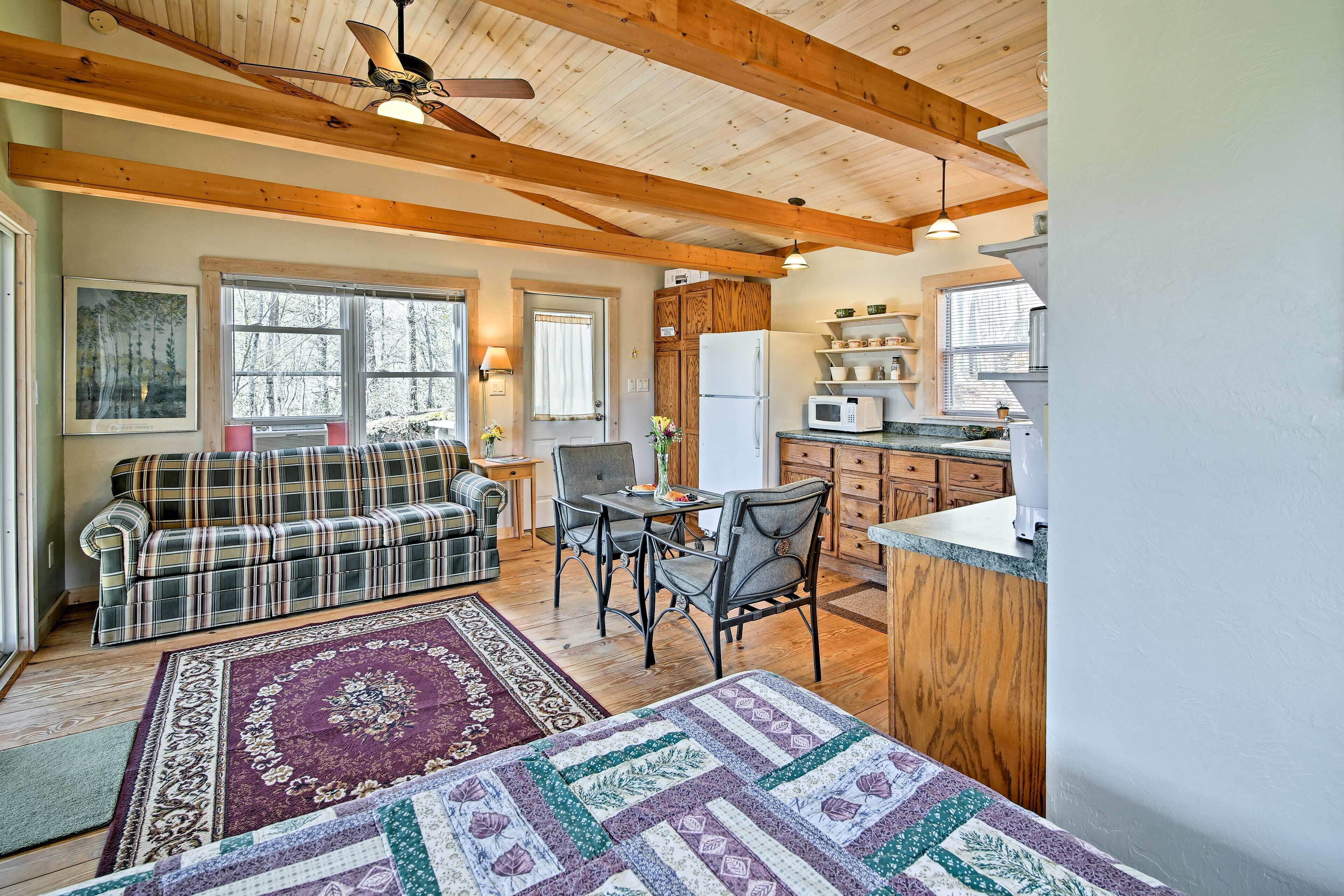 A cozy living room with a plaid sofa, a dining table with chairs, and a kitchen area with wooden cabinets and a microwave.