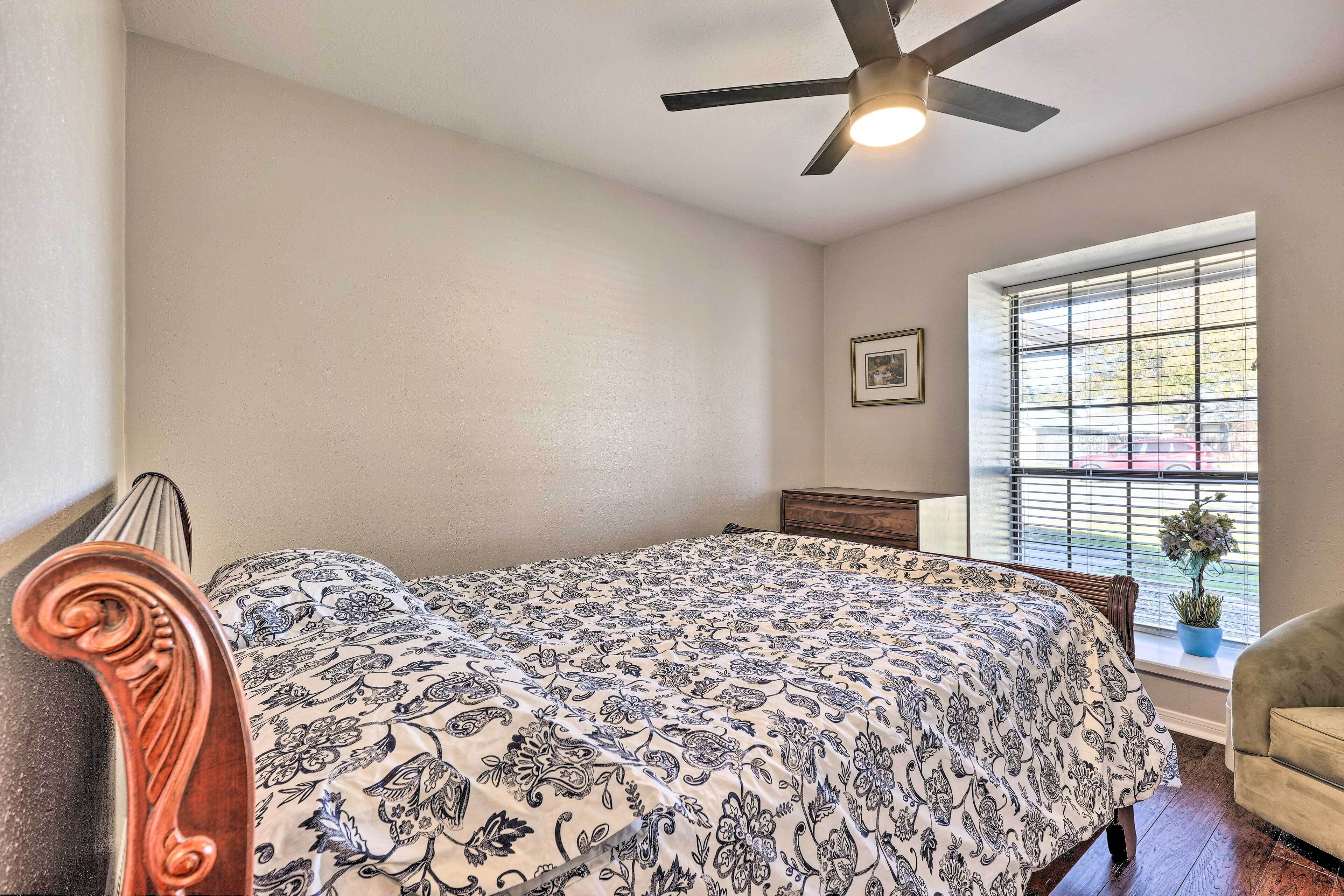 A bedroom with a bed, a ceiling fan, a window with blinds, a small table, and a chair.