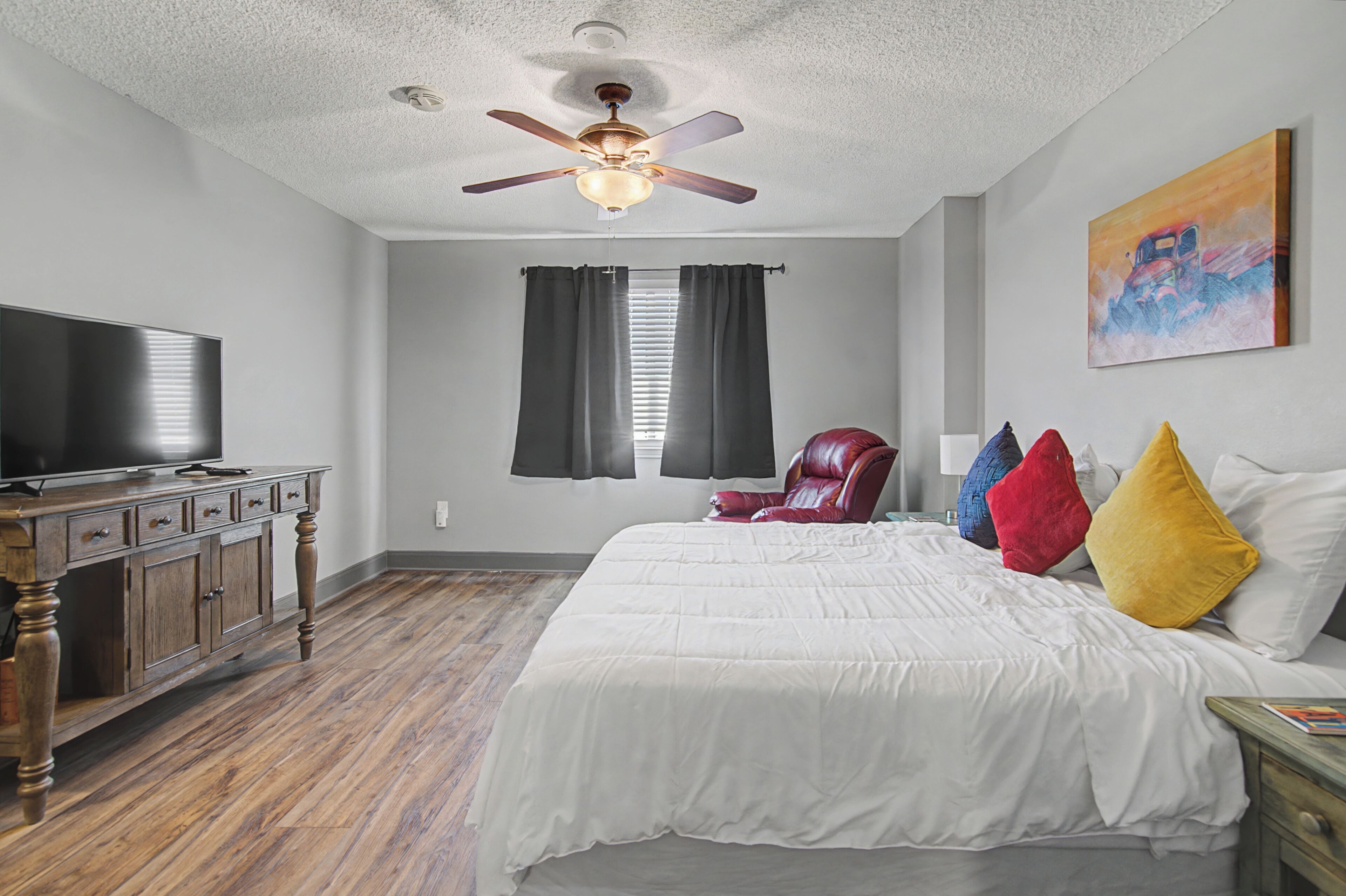 A bedroom with a bed, a TV, a ceiling fan, a window with curtains, and a painting on the wall.