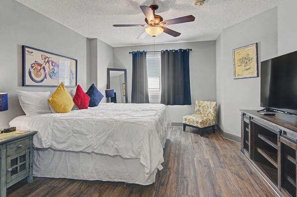 A hotel room with a bed, a chair, a TV, a ceiling fan, and artwork on the walls. - Shop & Dine: Fort Worth Condo w/ Racetrack Views! (Fort Worth)