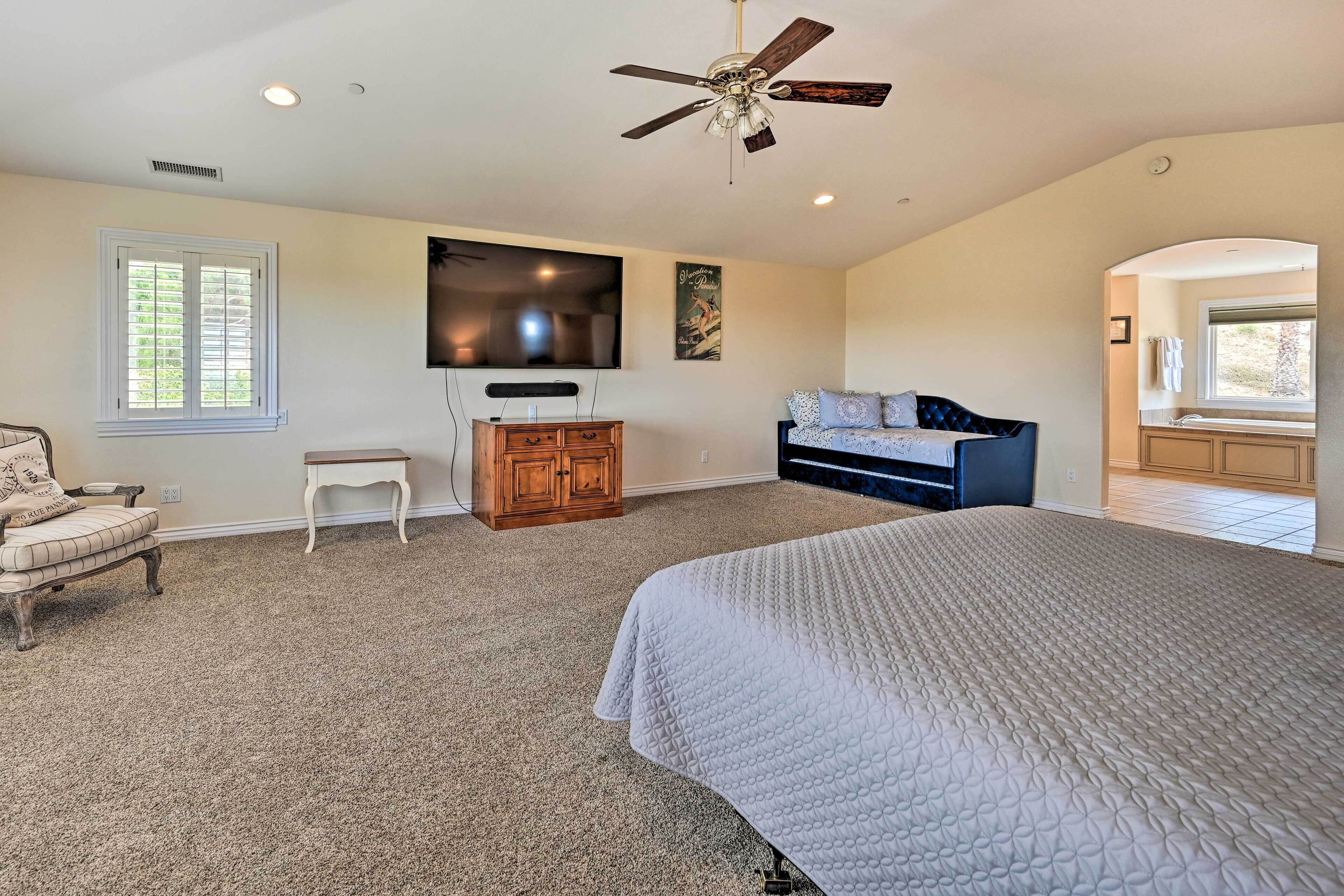 A bedroom with a large bed, a ceiling fan, a TV, a dresser, a chair, and a window.