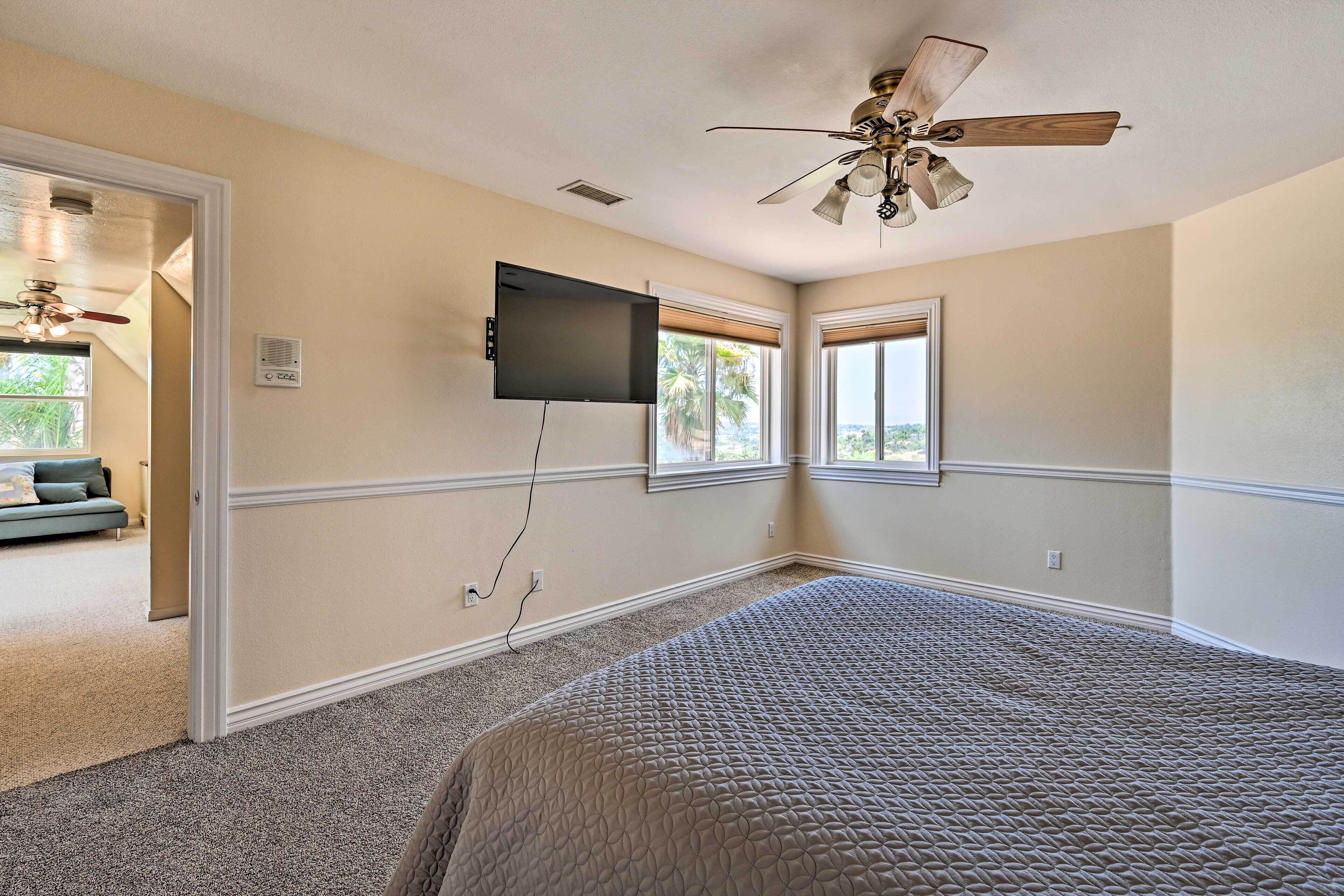 Un dormitorio con cama, ventilador de techo, televisor montado en la pared y dos ventanas.