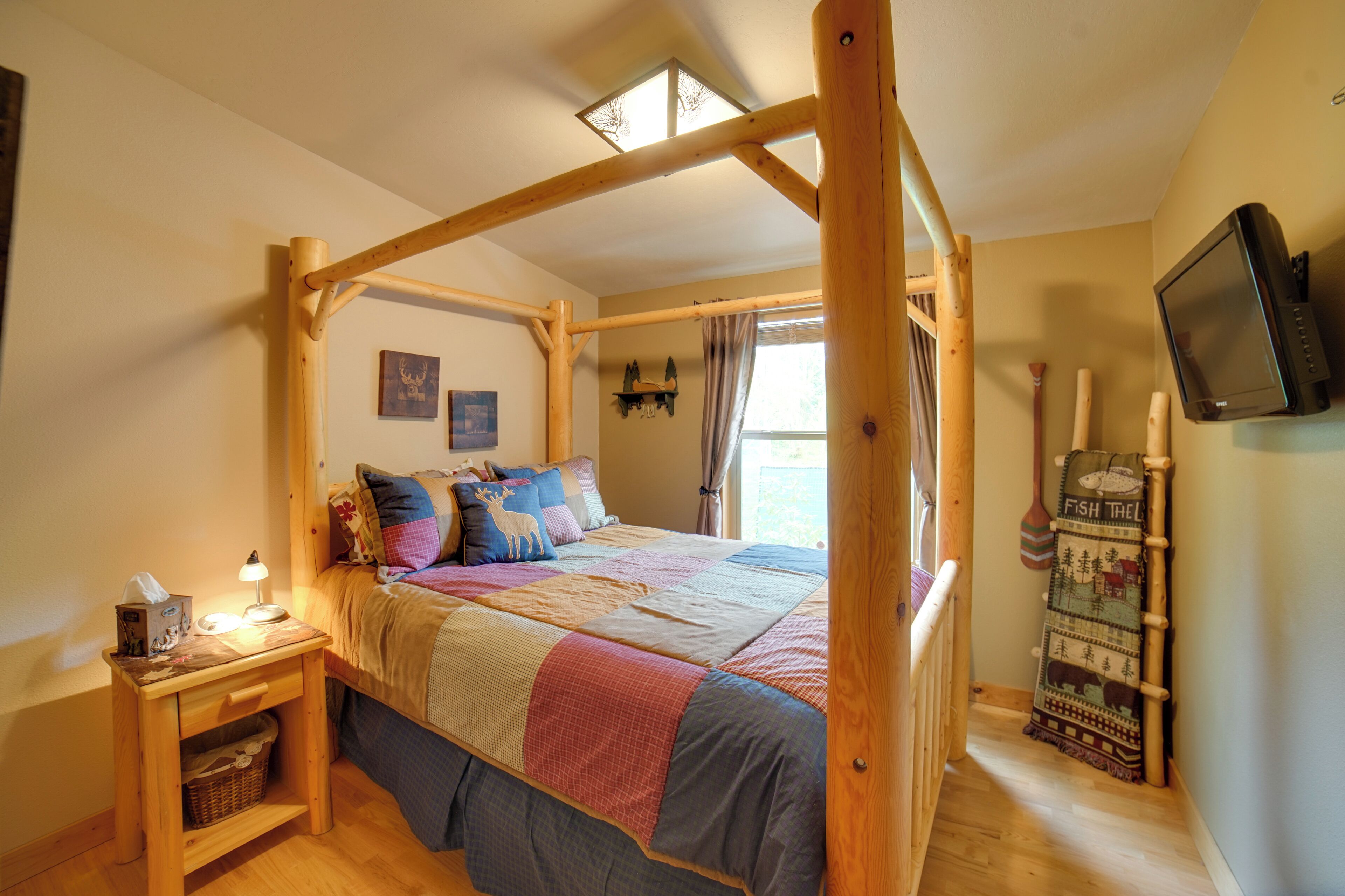 A bedroom with a wooden four-poster bed, a bedside table with a lamp, a television, and a wall-mounted shelf with decorative items.