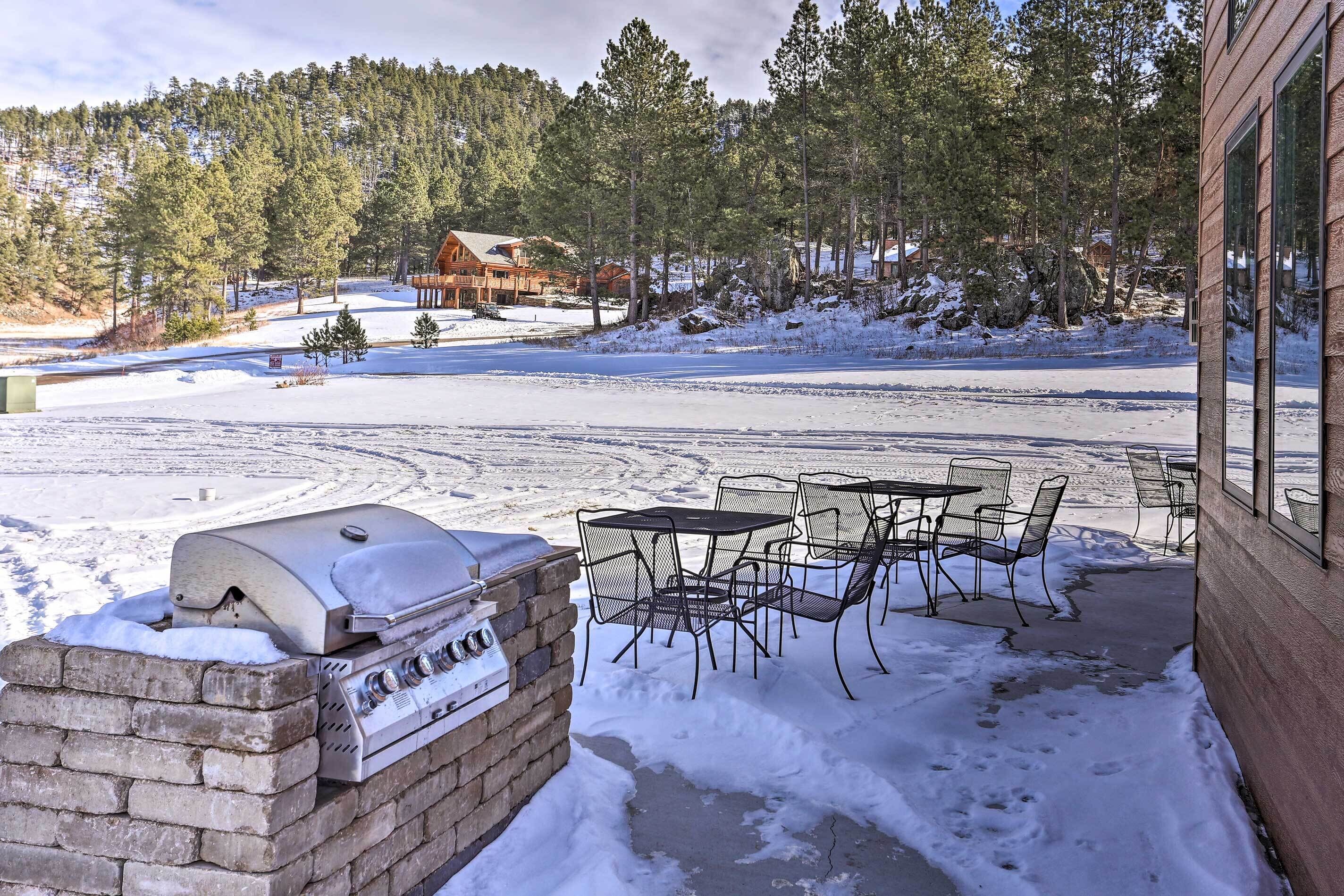 Une terrasse avec un barbecue, des sièges d’extérieur et une vue sur un chalet niché dans un paysage enneigé.