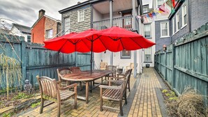 En terrasse med et træbord og stole, en rød parasol og en belagt sti af mursten.