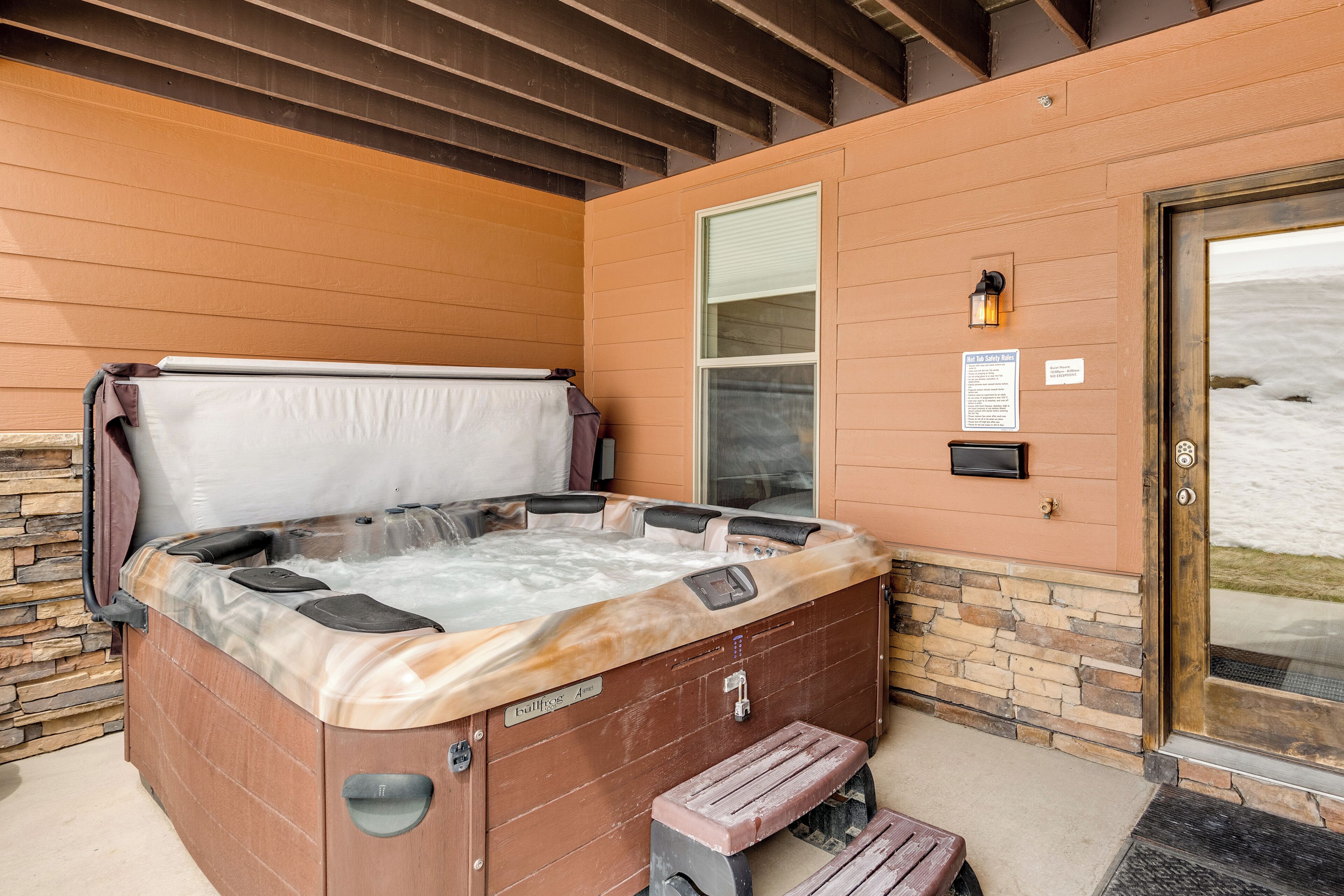 A hot tub with a stone wall backdrop, a wooden bench, and a door leading to a snowy landscape.