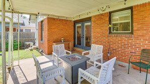 A patio with a fire pit table and white chairs, a brick wall, and a covered area with string lights.